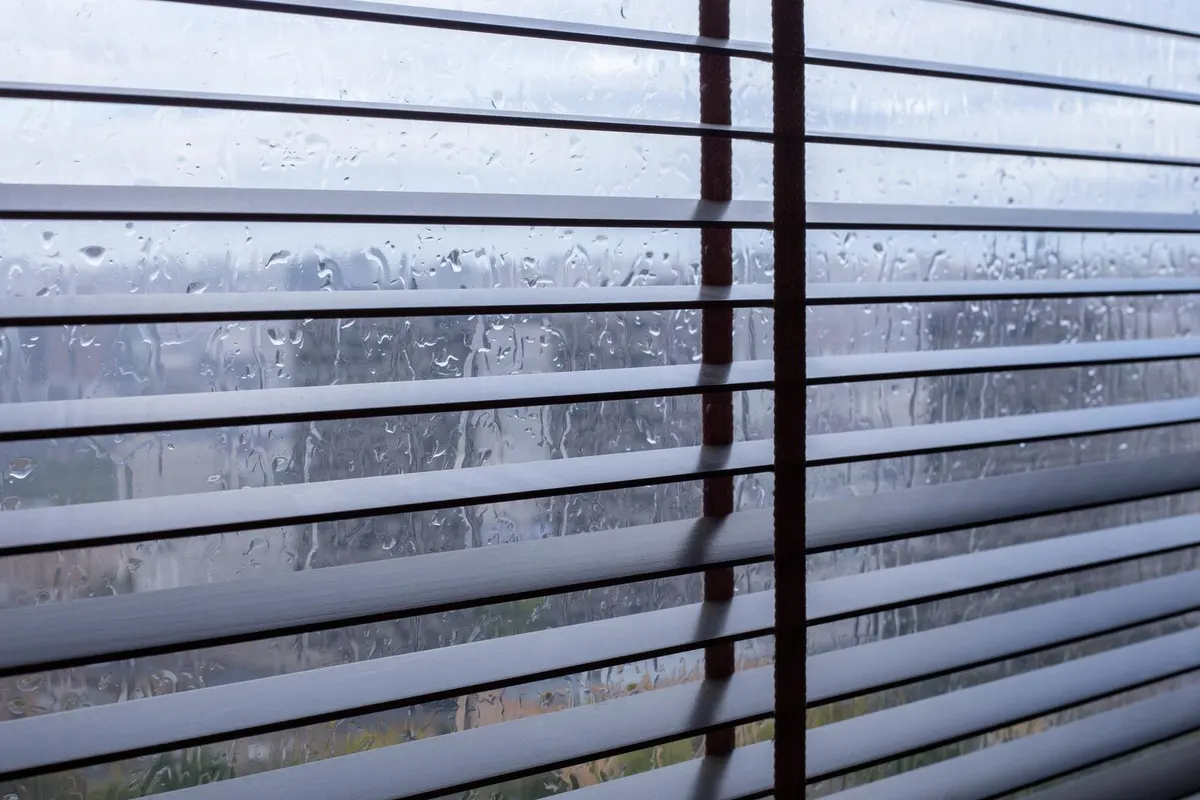 Lames d'un store vénitien intérieur derrière une vitre couverte de gouttes de pluie